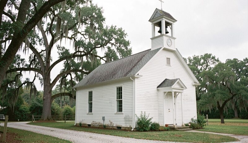 A small white clapboard country church with a little open belfry above its front gable, one dark bell hanging under a pointed roof, live oaks hung with Spanish moss beside a gravel lane.