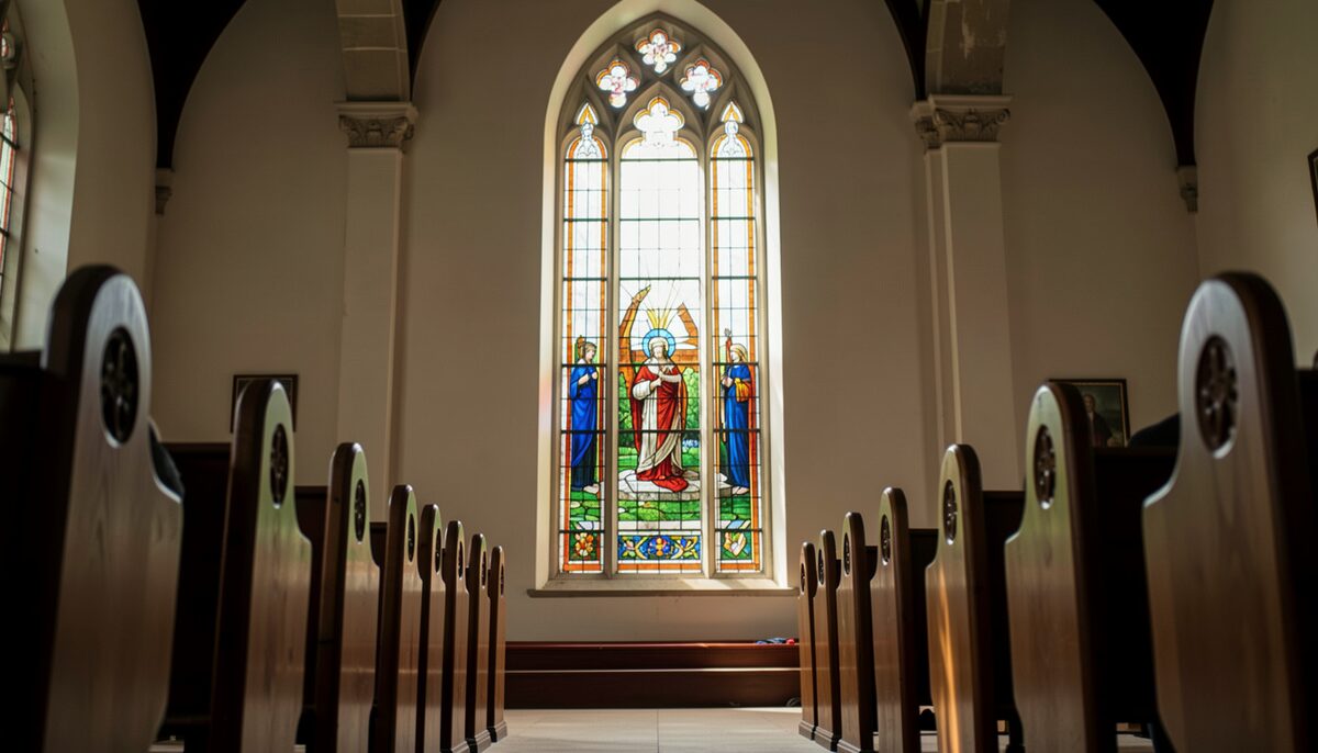 A tall stained glass window seen from inside, leaded panes glowing, pew backs in the foreground.
